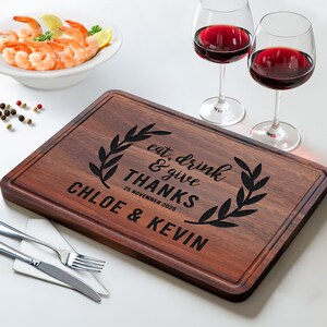 May include: A rectangular dark wood cutting board with a juice groove, engraved with a laurel wreath design and the text "eat, drink & give THANKS", "25 NOVEMBER 2025", and "CHLOE & KEVIN". Includes a fork and knife on a napkin, two wine glasses, and shrimp.