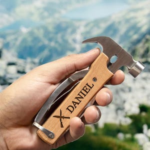 May include: A multi-tool with a wooden handle and a stainless steel hammer head. The handle is engraved with the name "DANIEL" and crossed knives. The tool also includes a folding knife and other implements.