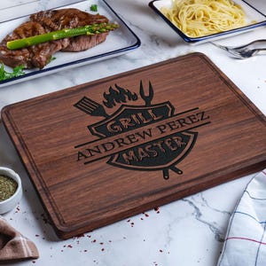 May include: A rectangular wooden cutting board with a dark brown finish. The board features a black engraved design with the words "GRILL MASTER" and the name "ANDREW PEREZ". The board is surrounded by plates of food.