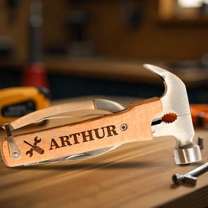 May include: A multi-tool hammer with a wooden handle engraved with the name "ARTHUR" and a wrench and screwdriver icon. The hammer features a claw, a knife, and other tools. The hammer is on a wooden surface.