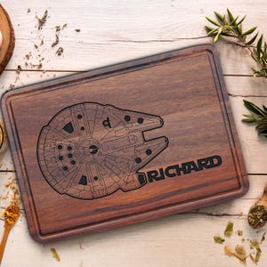 May include: A rectangular dark brown wooden cutting board featuring a laser-engraved outline of a spaceship and the name RICHARD. The board has a grooved perimeter channel and a smooth, polished surface with visible natural wood grain patterns.