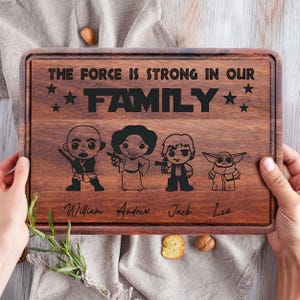 May include: A rectangular wooden cutting board with a dark brown finish. The board features engraved cartoon characters and the text "THE FORCE IS STRONG IN OUR FAMILY." Names are engraved below the characters. The board is held by hands.