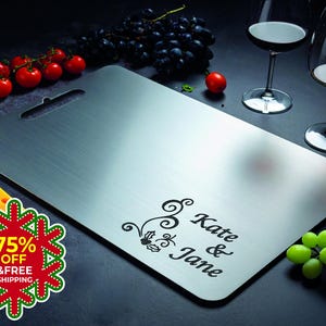 May include: A rectangular stainless steel cutting board with a handle, engraved with the names "Kate & Jane" and a decorative floral design. The board is surrounded by grapes, tomatoes, and wine glasses.