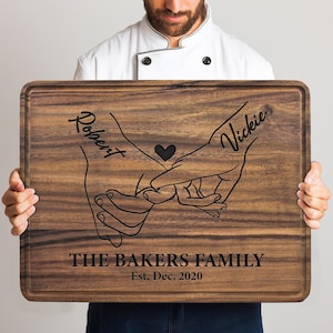 May include: A rectangular dark brown wooden cutting board featuring a laser-engraved design of two hands pinky promising with a small heart in the center. The names Robert and Vickie are engraved above, with The Bakers Family and Est. Dec, 2020 below.