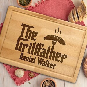 May include: A rectangular light-colored wooden cutting board with a juice groove along the perimeter. The board features a laser-engraved design with the text The Grillfather Daniel Walker and an illustration of a sausage on a fork.