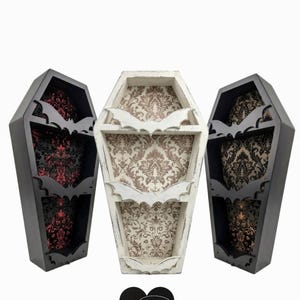 May include: Three coffin-shaped shelves in black and white, with bat cutouts and patterned interiors. The shelves have a distressed finish, and the interiors feature damask-style patterns in red, brown, and beige. The shelves are decorative.