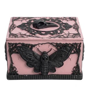 May include: A pink decorative box with black accents. The box features a black moth and raven skull design on the front and top. The box has a rectangular shape and is likely made of resin or a similar material.