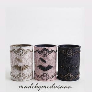 May include: Three cylindrical containers in white, pink, and black, each featuring ornate, raised baroque-style scrollwork and a central bat emblem. The text 'madebymedusaaa' is printed below the containers, followed by a small skull icon.