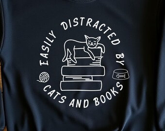 May include: Black sweatshirt with a white graphic design that says "Easily Distracted by Cats and Books". The graphic features a cat sleeping on a stack of books, a ball of yarn, and a bowl of cat food.