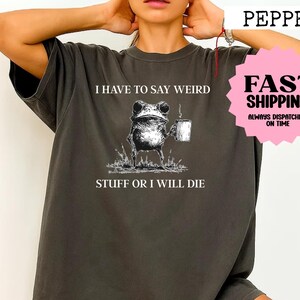 Puede incluir: Camiseta gris oscuro con una ilustración caprichosa de una rana sosteniendo una taza, con el texto "I HAVE TO SAY WEIRD STUFF OR I WILL DIE." La camiseta tiene un corte holgado y mangas cortas.