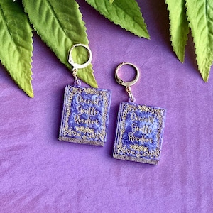 May include: A pair of earrings featuring miniature book pendants. Each purple book has gold lettering that reads "Dearest Gentle Readers." The earrings have gold-colored hoops and are set against a purple background.