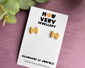 Può includere: Un paio di piccoli orecchini a forma di farfalla marrone chiaro su una carta bianca. La carta riporta il testo "HOW VERY JEWELLERY" e "HANDMADE IN NORFOLK @HowVeryJewellery". Lo sfondo è viola tenue con foglie verdi.