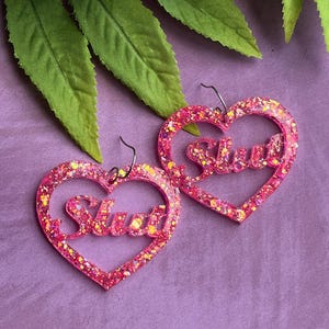 May include: A pair of pink heart-shaped earrings with glitter and the word "Slut" in cursive. The earrings are on a purple background with green leaves and white flowers.