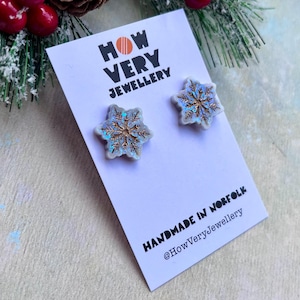 May include: A pair of snowflake stud earrings with a white background and gold glitter accents. The earrings are handmade in Norfolk and are sold by How Very Jewellery.