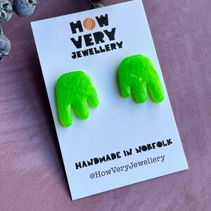 以下が含まれることがあります： ネオンの緑色の手型スタッドピアス。ピアスは「HOW VERY JEWELLERY」と「HANDMADE IN NORFOLK @HowVeryJewellery」の文字が入った白いカードに表示されています。
