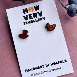May include: Squirrel-shaped stud earrings in shades of brown and red, displayed on a white card with the text "HOW VERY JEWELLERY" and "HANDMADE IN NORFOLK". The earrings are small, detailed, and appear to be made of wood. A sprig of berries is in the background.