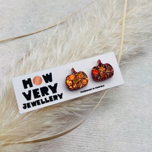 May include: A pair of small, glittery pumpkin-shaped earrings. The earrings are a deep burgundy colour with gold and orange glitter embedded in the resin. The earrings are on a white card with the text "HOW VERY JEWELLERY" and "HANDMADE IN NORFOLK".