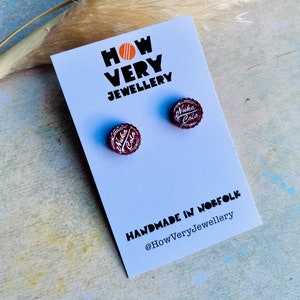 May include: Stud earrings with a Nuka Cola bottle cap design in red and white. The earrings are presented on a white card with the text "HOW VERY JEWELLERY" and "HANDMADE IN NORFOLK".