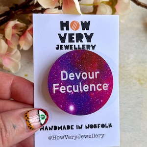 以下が含まれることがあります： 「Devour Feculence」の白い文字が入った、紫と赤の丸いボタン。ボタンは、「HOW VERY JEWELLERY」と「HANDMADE IN NORFOLK」の文字が入った白いカードにあります。