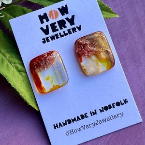May include: Square stud earrings with a distinctive design, showcasing amber, gold, and white tones. The earrings are presented on a white card with the text "HOW VERY JEWELLERY" and "HANDMADE IN NORFOLK".