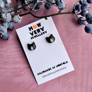 May include: Black cat stud earrings with white details, presented on a white card. The card features the text "HOW VERY JEWELLERY" and "HANDMADE IN NORFOLK @HowVeryJewellery". A sprig of blue berries is in the background.