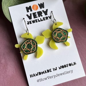 May include: Handmade turtle earrings. Each earring has a light green turtle shape with a glittery green and copper-coloured shell. The earrings are displayed on a white card with the text "HOW VERY JEWELLERY" and "HANDMADE IN NORFOLK".