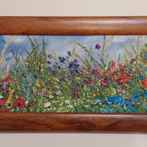 Puede incluir: Una pintura al óleo enmarcada que representa un campo de flores silvestres en tonos rojos, azules, morados y amarillos. La pintura está situada sobre un fondo de cielo azul con nubes blancas.