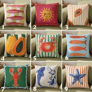 Peut inclure: Ensemble de coussins décoratifs avec divers motifs. Les coussins présentent des illustrations de poissons, d'un soleil et d'une lune, de papayes, d'un crabe, d'un homard, de carpes koï et d'une étoile de mer. Les coussins sont carrés et sont placés sur un canapé vert.