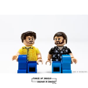 May include: Two Lego minifigures with brown hair and beards. The minifigure on the left is wearing a yellow shirt with a lightning bolt pattern and blue jeans. The minifigure on the right is wearing a black shirt with white stars and blue jeans.