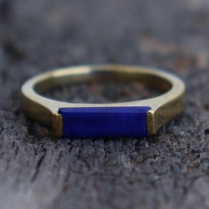May include: A gold-toned metal ring featuring a rectangular, deep blue lapis lazuli inlay set horizontally across the band. The ring has a smooth, polished finish and a flat, straight profile, showcasing a clean and simple design with no extra details.