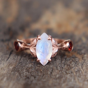 Peut inclure: Une bague en or rose avec une pierre de lune de forme marquise et deux petites pierres rouges de chaque côté. La bague est ornée d'un délicat motif de feuilles.