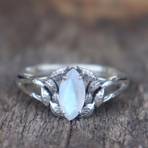 Könnte beinhalten: Ein silberner Ring mit einem tropfenförmigen, irisierenden Edelstein. Das Band des Rings weist ein detailliertes, blattartiges Design auf. Der Edelstein hat eine blassblaue Farbe und ist in eine dekorative Fassung gesetzt.