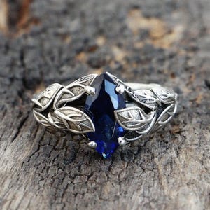 Könnte beinhalten: Ein silberner Ring mit einem tiefblauen, marquiseförmigen Edelstein. Der Ring hat ein Blattdesign um den Edelstein herum. Der Ring liegt auf einer verwitterten Holzoberfläche.