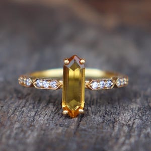 May include: A gold ring with a vertical, rectangular yellow gemstone and a brown tip. The band is decorated with small, clear gemstones. The ring is set against a blurred wooden background.