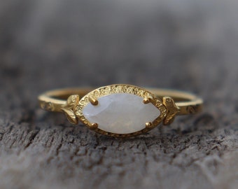 Puede incluir: Un anillo de oro con una piedra ovalada blanca engastada en una banda texturizada. El anillo tiene delicados detalles en forma de hojas a cada lado de la piedra.