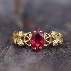 May include: A gold ring with a red gemstone set in a Celtic knot design. The ring is adorned with leaf details on either side of the stone.