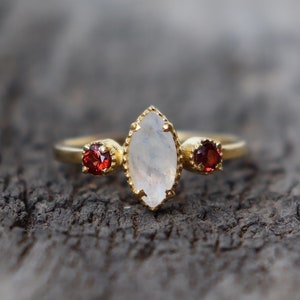May include: A gold ring with a white moonstone and two red gemstones. The ring is on a wooden surface.