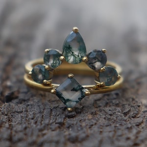 May include: A gold ring set featuring a band with a central square-cut moss agate stone and a curved nesting band adorned with five pear and round-cut moss agate stones, all set in gold prongs with visible green moss-like inclusions.