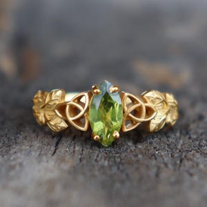 以下が含まれることがあります： 金色の指輪。中央にマーキス型の明るい緑色の宝石がセットされています。指輪のバンドには葉とケルトノットのデザインが施されています。素朴な木製の表面に置かれ、ジュエリーの繊細なディテールを際立たせています。