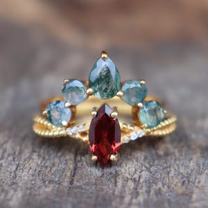 May include: A gold ring with a central, marquise-cut, deep red gemstone, surrounded by smaller, round, teal-colored stones. The ring has a delicate, textured band and is set against a blurred wooden background. A unique piece of jewelry.