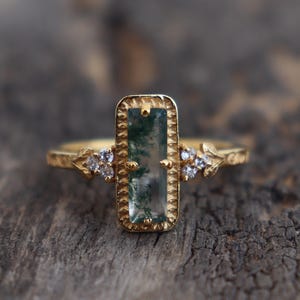 Può includere: Un anello d'oro con una pietra di agata verde rettangolare incastonata in una cornice dettagliata. L'anello è decorato con piccoli diamanti su entrambi i lati della pietra.