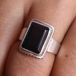 Puede incluir: Un anillo de plata con una piedra rectangular negra engastada en un engaste de plata. El anillo está en un dedo.