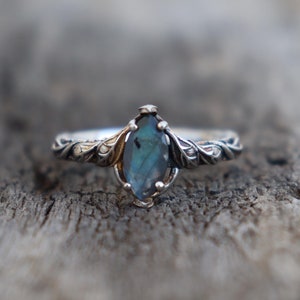 Pode incluir: Um anel de prata com uma pedra preciosa azul iridescente com corte marquesa. O anel apresenta um design ornamental em forma de folha ao redor da banda e da cravação. A pedra preciosa tem aproximadamente 1,3 cm de comprimento. O anel está sobre uma superfície de madeira rústica.