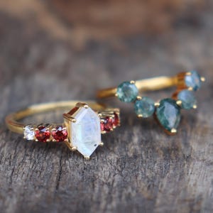May include: Two gold rings with gemstone accents. One ring features a large, multifaceted, light-colored stone flanked by smaller red and white stones. The other ring has a curved design with multiple blue-green gemstones.