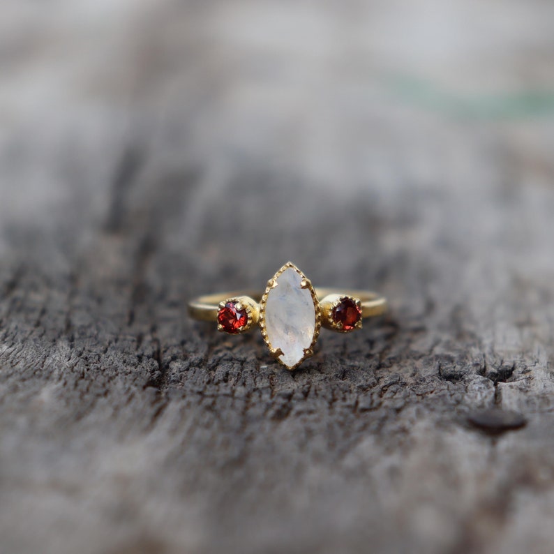 Może przedstawiać: Złoty pierścionek z białym kamieniem księżycowym i dwoma czerwonymi kamieniami szlachetnymi. Pierścionek leży na drewnianej powierzchni.