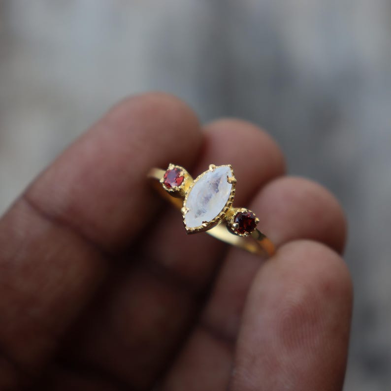 Może przedstawiać: Złoty pierścionek z dużym, białym, markizowym kamieniem szlachetnym i dwoma mniejszymi, czerwonymi kamieniami szlachetnymi po obu stronach.