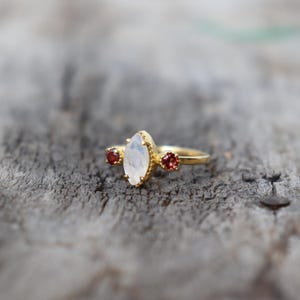 Może przedstawiać: Złoty pierścionek z dużym, owalnym kamieniem księżycowym i dwoma mniejszymi czerwonymi kamieniami po obu stronach.