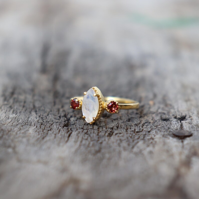 Może przedstawiać: Złoty pierścionek z dużym, owalnym kamieniem księżycowym i dwoma mniejszymi czerwonymi kamieniami po obu stronach.