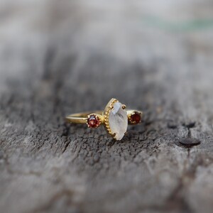 Może przedstawiać: Złoty pierścionek z białym kamieniem szlachetnym w kształcie markizy i dwoma mniejszymi czerwonymi kamieniami szlachetnymi po obu stronach.