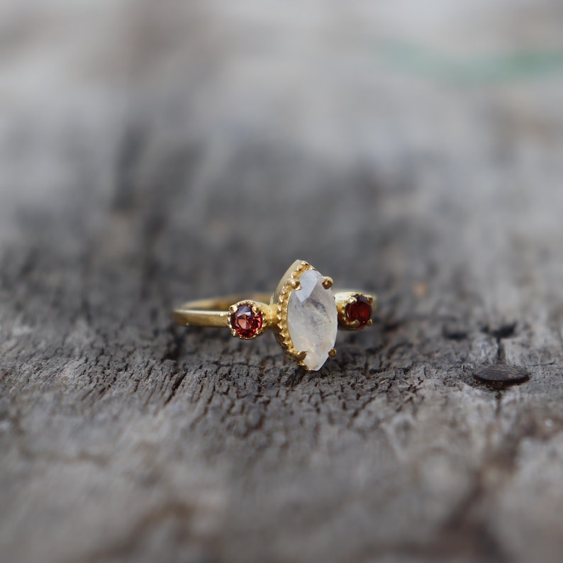 Może przedstawiać: Złoty pierścionek z białym kamieniem szlachetnym w kształcie markizy i dwoma mniejszymi czerwonymi kamieniami szlachetnymi po obu stronach.